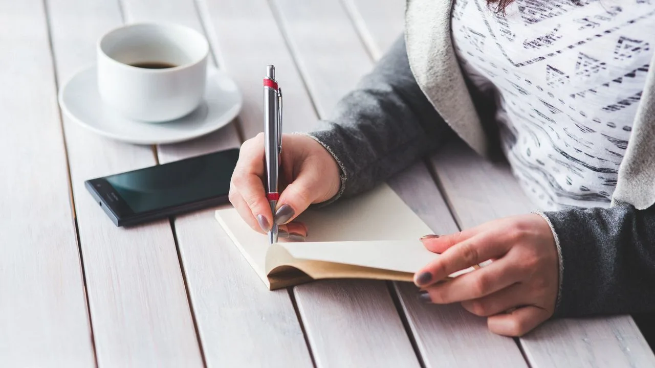 Mulher anota perguntas num caderno, ao lado de uma xícara de café, antes da consulta